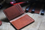 A brown Horween Dublin leather card case from the WM Leatherwork Collection by Tradevia is open on a wooden surface with various leather tools in the background, showcasing the beauty of a handcrafted leather piece.