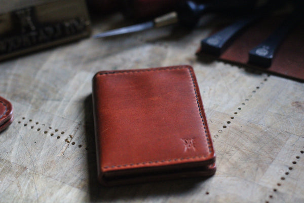 A WM Leatherwork Collection - Premium Leather Card Case by Tradevia, crafted from Horween Dublin leather, rests closed on a wooden surface, surrounded by leatherworking tools.