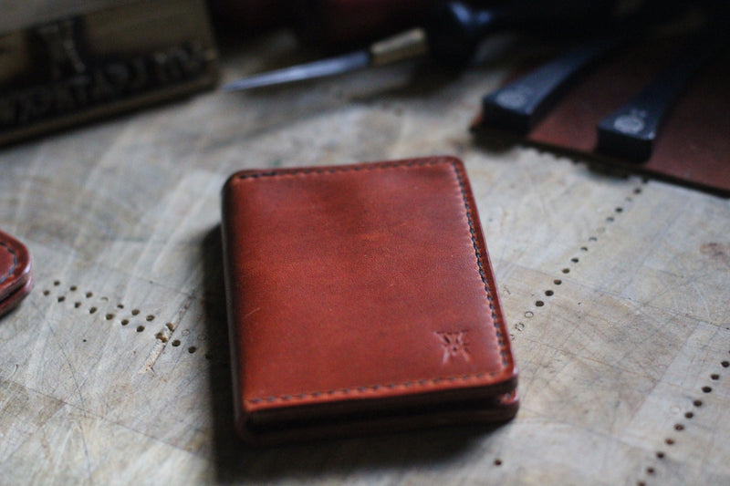 A WM Leatherwork Collection - Premium Leather Card Case by Tradevia, crafted from Horween Dublin leather, rests closed on a wooden surface, surrounded by leatherworking tools.