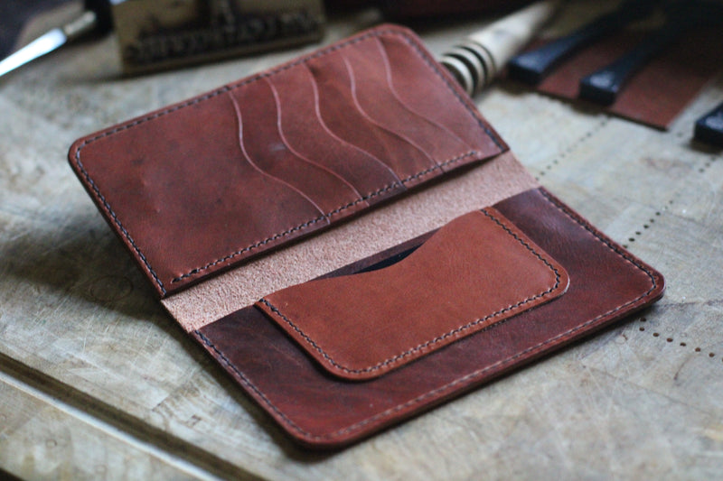 A WM Leatherwork Collection - Premium Leather Field Notes Wallet from Tradevia lies open on a wooden surface, showcasing several card slots and a pocket. Various leatherworking tools are scattered in the background.
