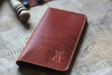 A WM Leatherwork Collection - Premium Leather Field Notes Wallet from Tradevia, crafted from brown leather with red stitching and an embossed 'W' logo, lies on a wooden surface next to a blurred object.