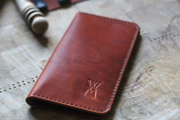 A WM Leatherwork Collection - Premium Leather Field Notes Wallet from Tradevia, crafted from brown leather with red stitching and an embossed 'W' logo, lies on a wooden surface next to a blurred object.