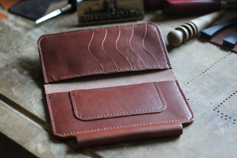 The WM Leatherwork Collection - Premium Leather Field Notes Wallet from Tradevia lies open on a wooden surface, revealing its multiple card slots and pocket. Various leatherworking tools are scattered in the background.