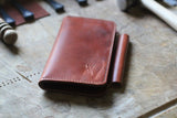 A brown leather Tradevia Wallet from the WM Leatherwork Collection, featuring a fold-over design and stamped logo, lies on a wooden surface surrounded by various tools.
