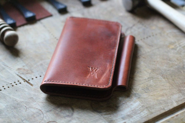 A brown leather Tradevia Wallet from the WM Leatherwork Collection, featuring a fold-over design and stamped logo, lies on a wooden surface surrounded by various tools.