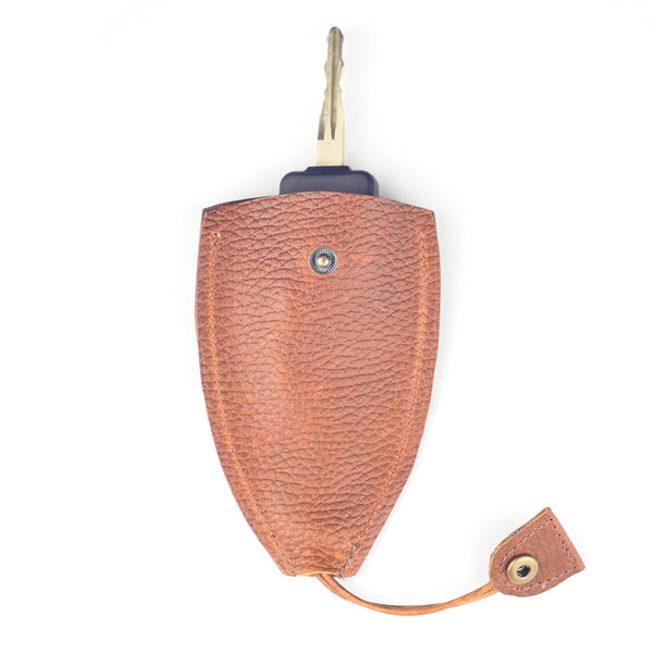 A single key in a brown textured leather key holder with a button clasp lies on a white background. This durable Tradevia Rogue Leather Key Protective Case ensures your keys are stylishly secured and easily accessible.