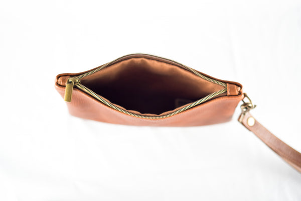 Top view of an open Sebago Leather Clutch by Tradevia, crafted from top-grain brown leather, featuring a prominent metal zipper and a detachable strap, set on a white surface. Made in the USA.