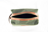 A green Waxed Canvas Dopp Kit by Tradevia with a brown leather zipper partially open, viewed from above. Made in the USA.