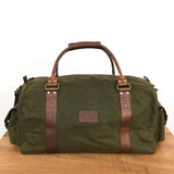 A Tradevia White Cap Waxed Canvas Duffle with full-grain leather straps and handles is placed on a wooden surface against a plain white background.