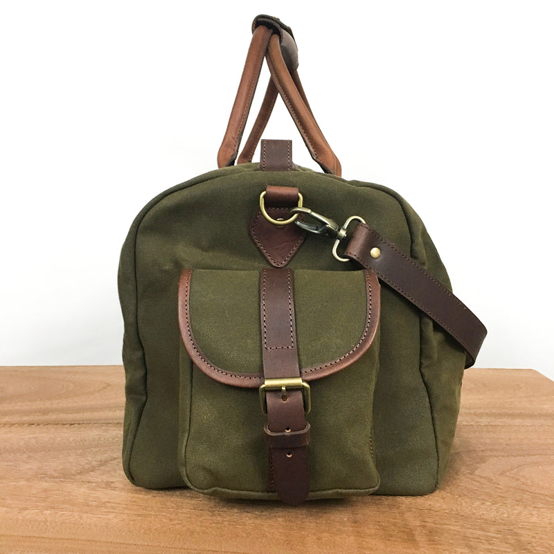 A White Cap Waxed Canvas Duffle from Tradevia in green, featuring brown leather accents, a front pocket with a buckle closure, and a full-grain leather shoulder strap, sits on a wooden surface against a plain white background. The water-resistant waxed canvas ensures durability and style.