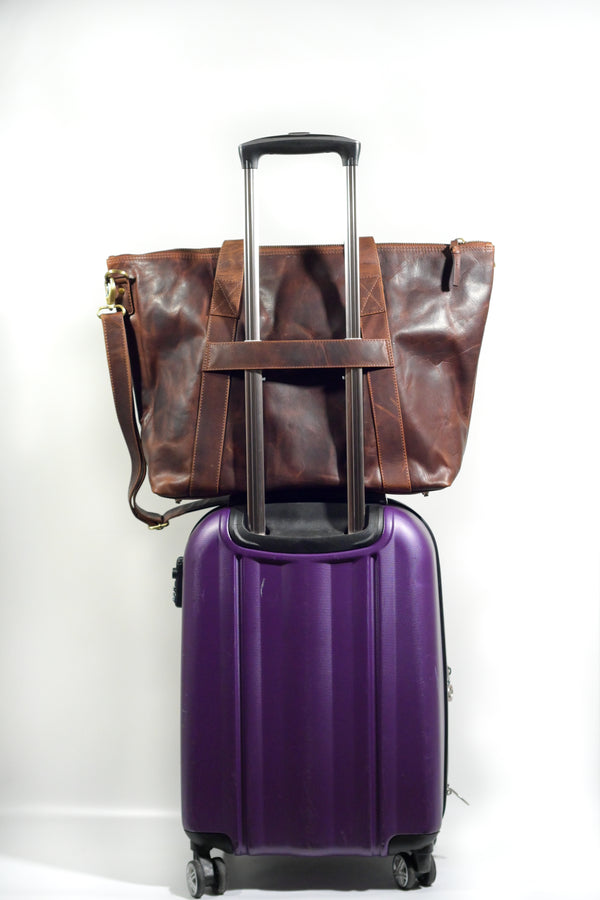 A purple suitcase with a luggage strap on top of a brown Tradevia Rockport Leather Weekend Tote.