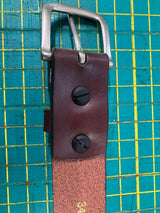 Close-up of a handcrafted Baxter Belt and Made in Maine Rogue Wallet Bundle by Tradevia with a silver buckle, featuring black screws securing the buckle. A green cutting mat with yellow grid lines serves as the background.