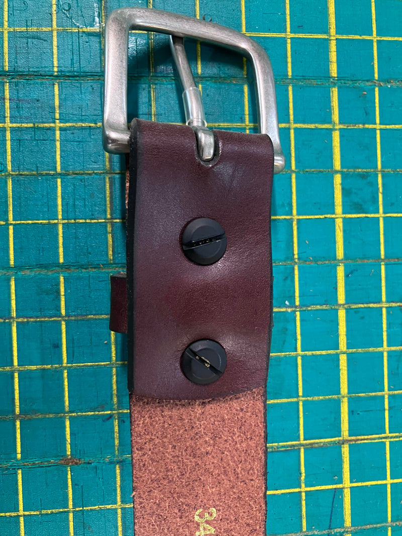 Close-up of a handcrafted Baxter Belt and Made in Maine Rogue Wallet Bundle by Tradevia with a silver buckle, featuring black screws securing the buckle. A green cutting mat with yellow grid lines serves as the background.