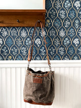 A Tradevia Kennebunkport Bucket Bag with an adjustable shoulder strap is hanging on a wall next to a blue wallpaper.