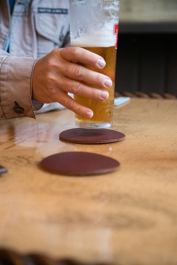 A person drinking a beer on a wooden table with Sebago Camp Coasters coasters by Tradevia.