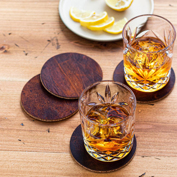 Two glasses of whiskey on a wooden table, resting on Tradevia Genuine Moose Leather Coaster Set.