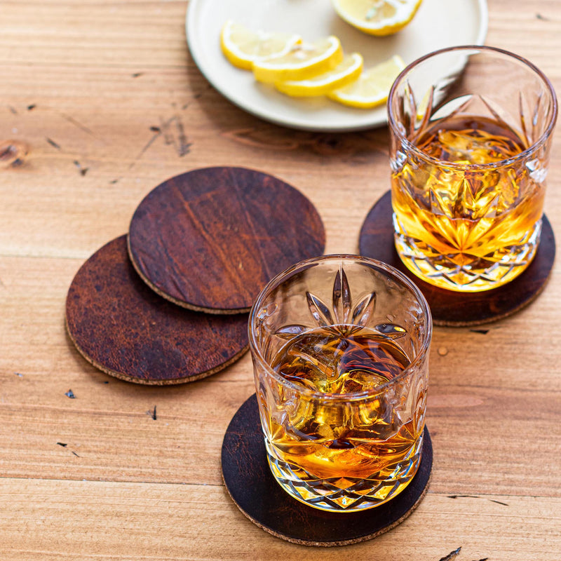 Two glasses of whiskey on a wooden table, resting on Tradevia Genuine Moose Leather Coaster Set.