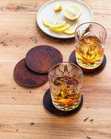 Two glasses of whiskey on a wooden table, resting on a Tradevia Moose Leather Coaster Set.