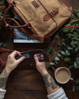 A person holding a Tradevia Waxed Canvas Laptop Bag next to a durable wallet and a cup of coffee.
