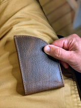 A hand is holding a slim Factory Second PREVIOUSLY EMBOSSED Made in Maine Front Pocket Wallet - BROWN by Tradevia against a background of khaki-colored pants. This stylish accessory, crafted in Maine, features RFID-blocking technology for added security.