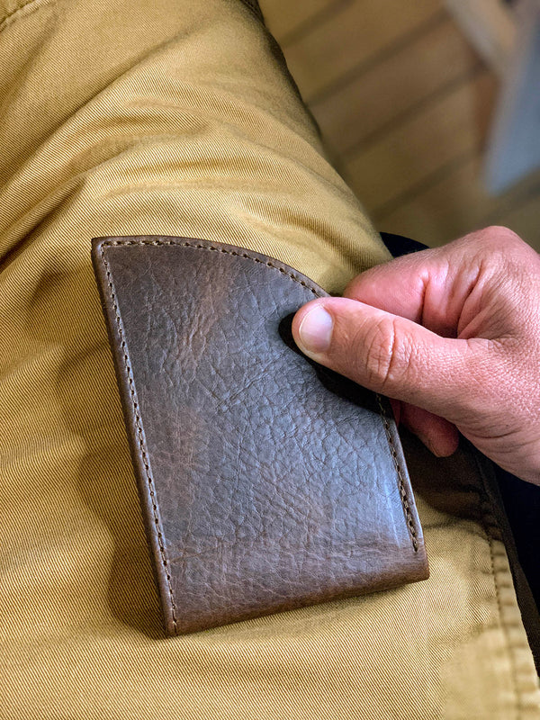 A hand is holding a slim Factory Second PREVIOUSLY EMBOSSED Made in Maine Front Pocket Wallet - BROWN by Tradevia against a background of khaki-colored pants. This stylish accessory, crafted in Maine, features RFID-blocking technology for added security.