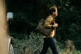 A man walking in the woods, carrying a durable Tradevia Waxed Canvas Tote Bag with top-grain leather straps.